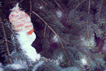 cheerful snowman in hat hiding in the shade of a tree from sun / warming smile to a good weekendの写真素材