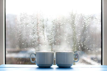 couple cup coffee with a frothy cappuccino in the background window with raindrops / cozy warming morningの写真素材