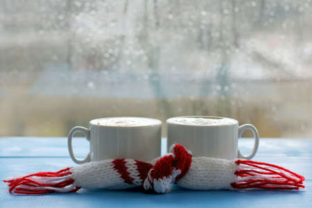 two cups of frothy cappuccino with a scarf on the background of the window, with drops after the rain / warming atmosphere of spring coffee breakの写真素材