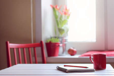 Red mug with a hot drink and a paper notebook on the window background / working in a cozy home environmentの写真素材