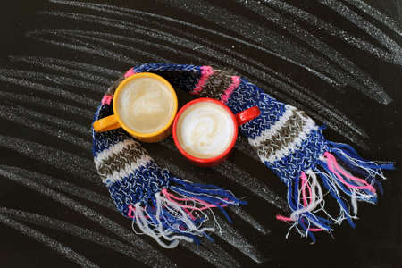 two mugs frothy cappuccino with scarf wrapped on background of chalkboard top view/ warming coffee breakの写真素材