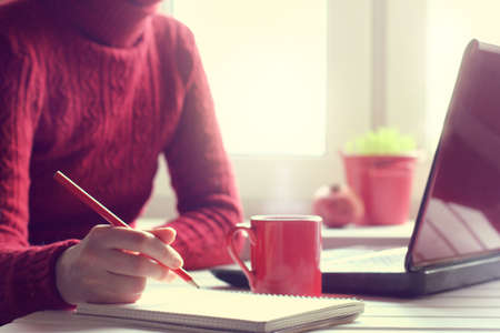 man in red sweater makes notes in pencil on the background of working environment / atmosphere cozy workの写真素材