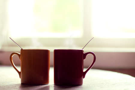 Red and yellow mugs with a hot drink on the table by the window / Breakfast together in a cozy atmosphereの写真素材
