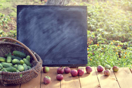 Old wicker basket with a cucumbers  crop on a table with apples and a board for graffiti/ vegetables and fruitsの写真素材