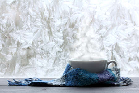white mug in a blue scarf and warm steam in the form of a Christmas tree against the background of a frozen window/ warming Christmas fantasyの写真素材