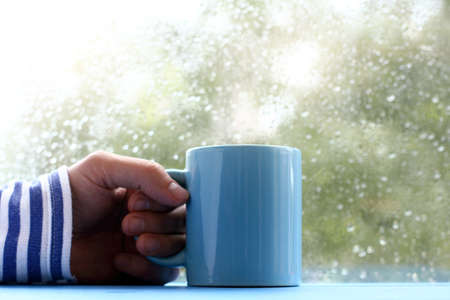 hand in striped sweater with and blue cup on table, against the window with drops after rain / autumn mood in a marine styleの写真素材