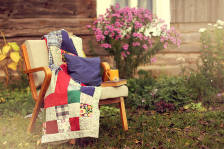 old chair with a blanket, a book and a yellow mug stands in the garden near the wooden house / cozy...