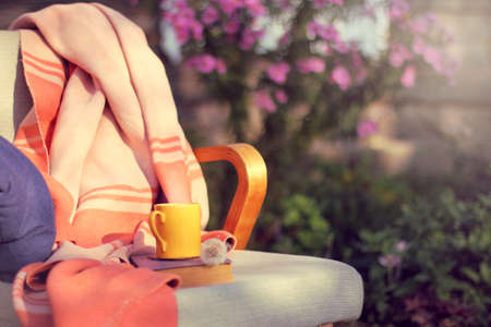 Armchair  with  blanket, pillow, book and cup on the background of  flower bed in the garden / warming atmosphere for relaxationの写真素材