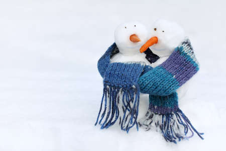 two smiling snowmen with carrot noses in a blue scarf on a background of a snowy landscape / warming the pastime of a winter vacationの写真素材