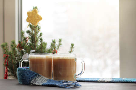 two glass glasses with frothy cappuccino on the background of festive Christmas tree by the window / winter coffee breakの写真素材