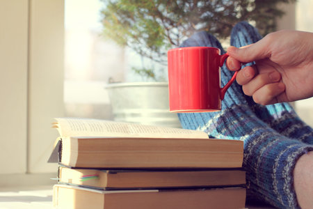 feet in warm socks and a hand with a red mug on the table with books on the background of a window...