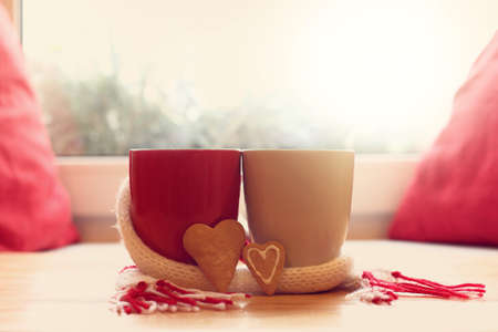 two mugs wrapped in scarf next to pair of cookies in the shape of heart on table in front of window / together in cozy atmosphereの写真素材