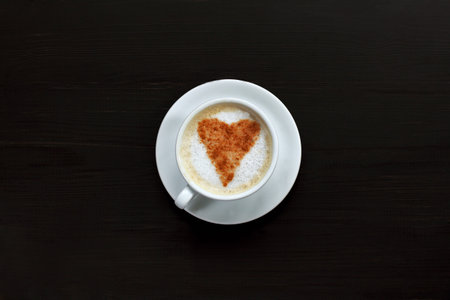 cup of frothy cappuccino decorated with heart of cinnamon stands on dark table top view / hot love with the aroma of coffeeの写真素材