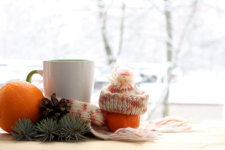 cup in a scarf and a mandarin in a hat decorated with a fir branch and a cone against the window....
