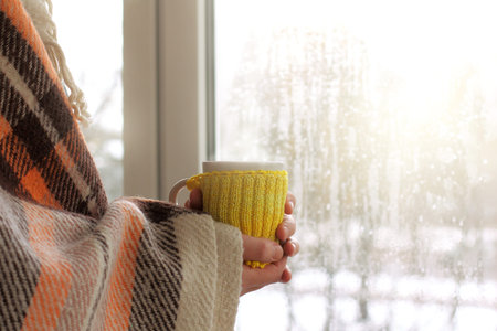mug in a yellow sweater in the hands wrapped in a plaid against the window after the first rain /...