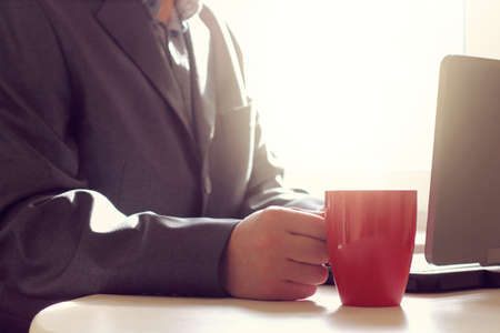 red cup in the hand of the manager sitting at the table with a laptop on the background of the morning window. warming work environmentの写真素材
