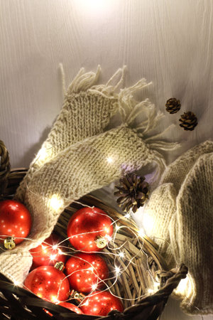 red Christmas balls in a basket with a scarf and mittens and cones on the table in the evening....