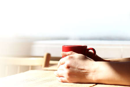 red mug in hands on the background of a window with a copy space. all you need for a warm morningの写真素材