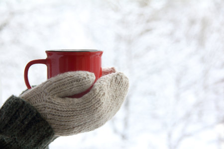 a red mug in the hands of those dressed in knitted mittens against the backdrop of a blurred snow landscape. a warming drink for a winter morningの写真素材