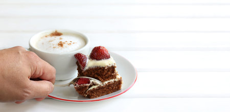 hand with a spoon separates a piece of chocolate and strawberry cake on the background of a cup with...