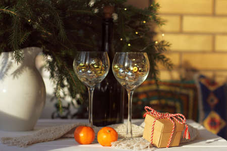 gift with a striped bow and tangerines with a scarf on the table on the background of wine glasses and a Christmas tree with a garland. winter holiday tableの写真素材