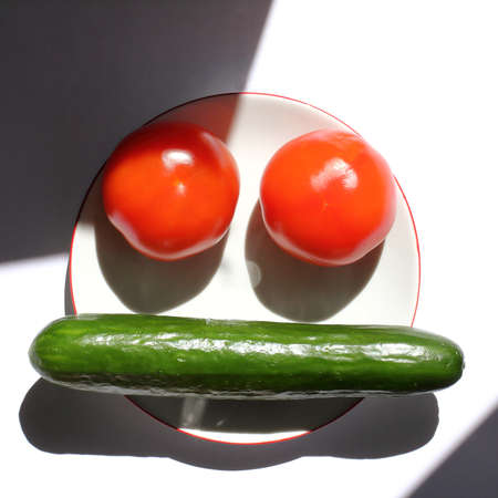 two red tomatoes and a green cucumber on a plate on a sunny day with shadows. funny vegetable avatarの写真素材