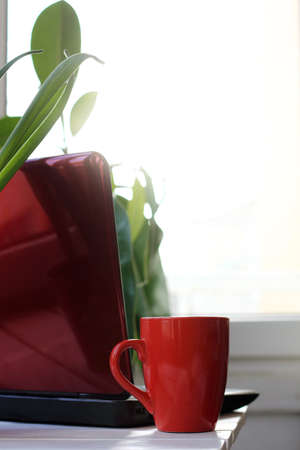 red mug and laptop on the table against the background of the morning window. warming drink before starting workの写真素材