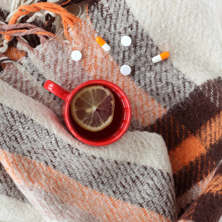 tea with lemon in a red mug wrapped in a blue scarf on a table with a thermometer and pills top...
