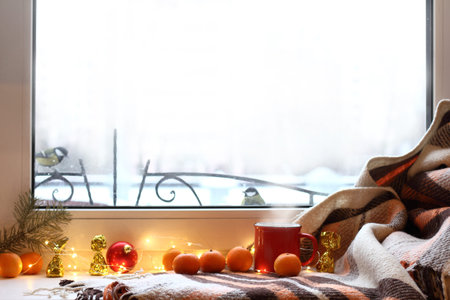 red mug of hot drink on a windowsill with a warm blanket and tangerines when behind is window...