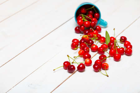 red cherries scattered from the stylish blue cup on a white wooden backgroundの写真素材