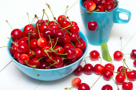 red cherries scattered from the stylish blue cup on a white wooden backgroundの写真素材
