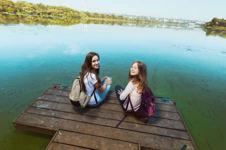 Two teenager smiling girls with backpacks sitting on a pier at the river bank and the city in the backgroundの写真素材