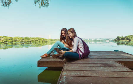 Two teenager smiling girls with backpacks sitting on a pier at the river bank and the city in the backgroundの写真素材