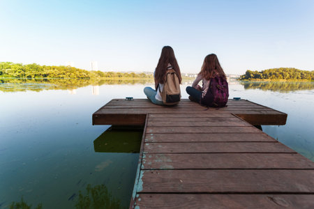 Two teenager girls with backpacks sitting on a pier at the river bank and the city in the background