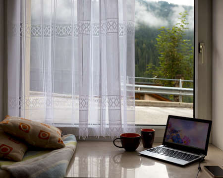 wonderful morning in the mountains. pillows, plaid, laptop, cup of coffee on the windowsill and the landscape outside the windowの写真素材