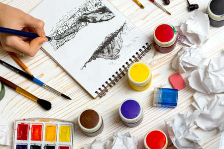 girl paints the landscape in graphics technics. the hand of the girl artist and Album pages, paints, pencils,oil, brush on a wooden backgroundの写真素材