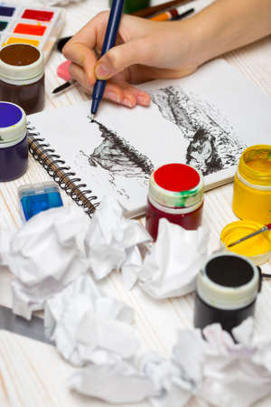 girl paints the landscape in graphics technics. the hand of the girl artist and Album pages, paints, pencils,oil, brush on a wooden backgroundの写真素材