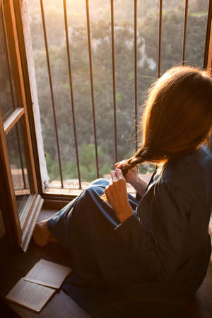 girl is sitting on the floor by the open window at the sunset and tidies the hairの写真素材