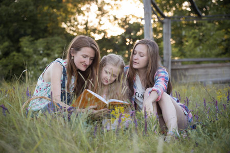 Happy family - mom and two daughters are sitting in a meadow and reading a bookの写真素材
