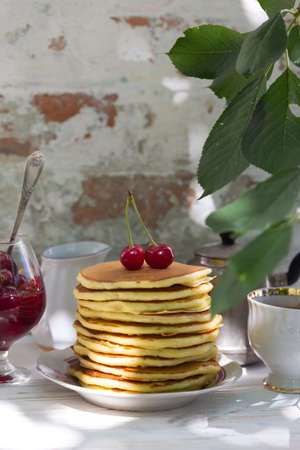 Pancakes and cherry jam stand on the table with a cup and a brewer on the background of a vintage brick wallの写真素材