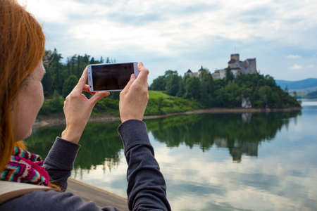 girl taking a picture of a beautiful castle on a lake shore with her smartphoneの写真素材