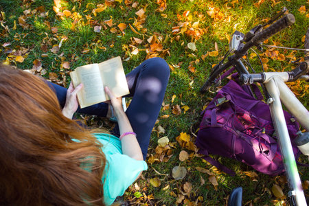 girl in the park sits under a tree and reads a book. next to it there is a bicycle and a backpackの写真素材