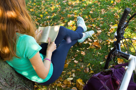 girl in the park sits under a tree and reads a book. next to it there is a bicycle and a backpackの写真素材