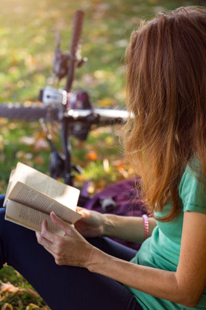 girl in the park sits under a tree and reads a book. next to it there is a bicycle and a backpackの写真素材