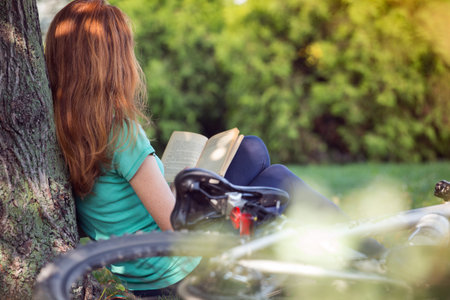 girl in the park sits under a tree and reads a book. next to it there is a bicycle and a backpackの写真素材