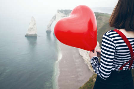Happy girl with a red balloon in the shape of a heart at background of scenery Etretat. northern coast of Franceの写真素材