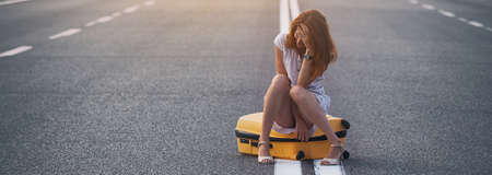 problems in the journey. the girl is sitting on a yellow suitcase on the dividing strip on the road.の写真素材