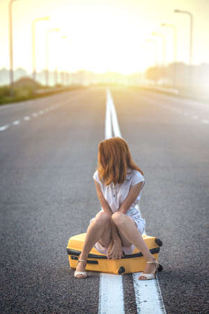 problems in the journey. the girl is sitting on a yellow suitcase on the dividing strip on the road.の写真素材