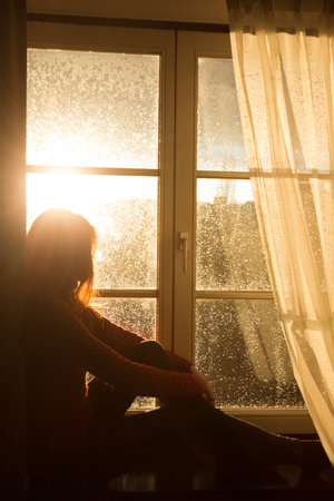 girl sitting on a windowsill and looking at the evening city at the beams of orange sunset sunの写真素材