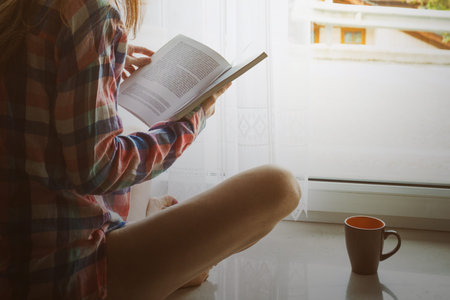 girl holding a book sitting on a windowsill with bright sun light from the windowの写真素材